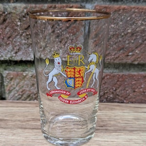 Op de afbeelding: Een helder glas met een gouden rand, met een kleurrijk embleem met een kroon, leeuw, eenhoorn en de tekst "Coronation of Queen Elizabeth II June 2nd 1953". Het glas staat tegen een bakstenen achtergrond.