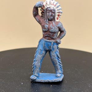May include: A small, vintage toy figurine of a Native American chief. The figure is painted with brown skin, blue trousers, and a white and red headdress. The figure is standing on a blue base.