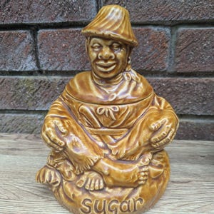 May include: A brown ceramic sugar jar shaped like a seated figure wearing a hat. The figure has a brown face and is holding its legs. The jar has the word "Sugar" written on the bottom.