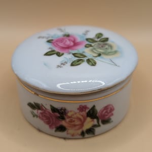 May include: A white porcelain trinket box with a gold trim and floral designs. The lid features a pink, white, and yellow rose with green leaves. The base of the box has a similar floral design with pink, yellow, and white roses.