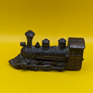 May include: A small, antique-style train engine figurine in a bronze colour. The miniature locomotive features detailed elements such as a smokestack, cowcatcher, and windows. The train is set against a bright yellow background.