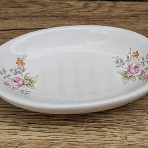 May include: White oval soap dish with a floral design. The dish features a raised rim and three ridges for drainage. The floral pattern includes pink roses, blue and orange flowers, and green leaves. The dish is made of ceramic.