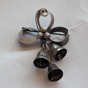Puede incluir: Un broche de plata con un diseño de lazo estilizado y tres campanas colgantes. El lazo está hecho de alambre retorcido y las campanas están unidas a una cadena.