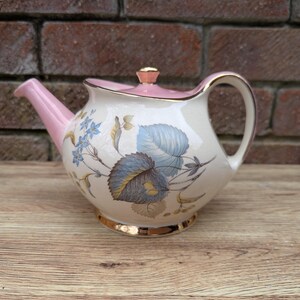 May include: A vintage teapot with a pink lid and a floral design featuring blue and yellow leaves on a white background. The teapot has a gold trim around the base.