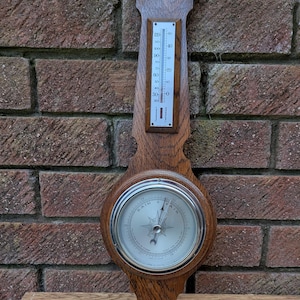 May include: A vintage wooden barometer and thermometer combination. The barometer features a silver-colored dial with a needle and the words "Rain" and "Fair." The thermometer displays Fahrenheit and Centigrade scales. The wooden frame has a dark brown finish.