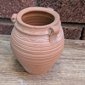 Puede incluir: Una vasija de arcilla color terracota con una amplia abertura circular, dos pequeñas asas laterales y una serie de estrías horizontales y patrones de líneas onduladas grabados en la superficie exterior del cuerpo.