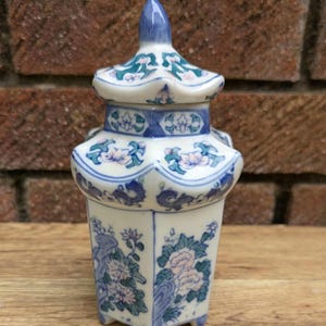 Pode incluir: Um pequeno recipiente de porcelana ornamentado com tampa. O recipiente é branco com padrões e detalhes florais azuis e rosa. A tampa é encimada por um pináculo azul. O recipiente tem formato de pagode.