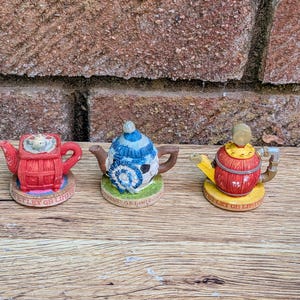 Puede incluir: Tres teteras decorativas en miniatura con diseños únicos. La izquierda es roja con base cuadrada, la central es azul y blanca con rayas, y la derecha es roja y amarilla con forma de barril. Cada base tiene la inscripción Tetley GB Limited.