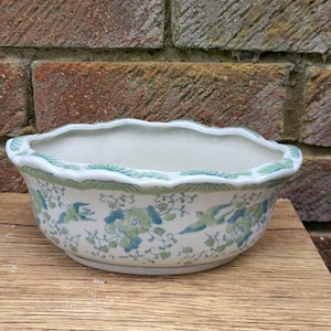 May include: A white ceramic planter with a scalloped rim, adorned with blue and green floral and bird patterns. The planter sits on a wooden surface, showcasing a classic, decorative design.