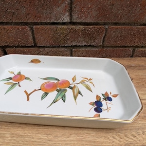 May include: A rectangular white ceramic serving dish with a gold rim. The dish is decorated with a colourful illustration of peaches and blueberries on branches with green leaves. The dish is on a wooden surface.