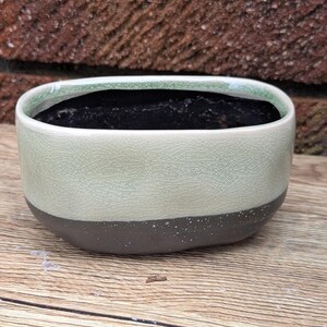 May include: An oval ceramic planter featuring a two-tone design with a light green crackle glaze on the upper portion and a dark brown matte finish on the lower base. The interior is dark and hollow, designed for holding small plants or succulents.