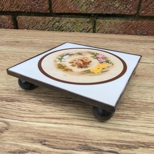 Peut inclure: Un dessous de plat carré avec une surface blanche et une bordure marron. Le centre présente une illustration détaillée d'un oiseau entouré de fleurs. Le dessous de plat est soutenu par quatre petits pieds sphériques foncés. Un article décoratif pour la cuisine.