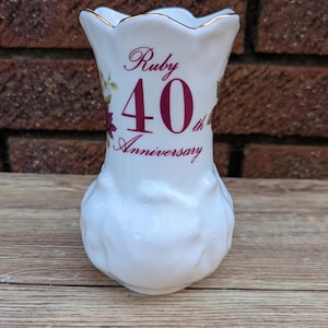 Può includere: Un vaso in ceramica bianca con la parte superiore svasata e la base arrotondata. Il vaso è decorato con le parole "Ruby 40th Anniversary" in un carattere corsivo, con accenti floreali. Il bordo è rifinito con oro.