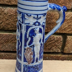 May include: A blue and white ceramic beer stein featuring a raised design of a knight in armour and a shield. The stein has a handle shaped like a dragon's head.