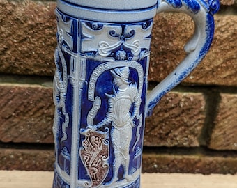 Puede incluir: Una jarra de cerveza de cerámica azul y blanca con un diseño en relieve de un caballero con armadura y un escudo. La jarra tiene un asa con forma de cabeza de dragón.
