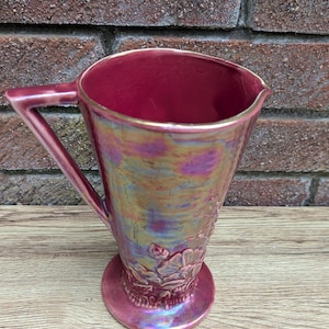 Może przedstawiać: Bordowy dzbanek z trójkątnym uchwytem i rozszerzanym korpusem. Dzbanek ma metaliczne, opalizujące wykończenie z nutami złota i fioletu. Podstawa ma kwiatowy wzór reliefowy. Dzbanek jest prawdopodobnie ceramiczny lub szklany.