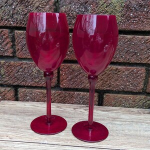 May include: Two matching claret glasses in a rich burgundy colour. The glasses feature a traditional design with a rounded bowl, a thin stem, and a circular base. They are set on a wooden surface against a brick wall background.