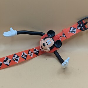 Könnte beinhalten: Eine rote Mickey Mouse-Uhr mit einem schwarzen Armband mit einem sich wiederholenden Muster aus schwarz-weißen geometrischen Formen. Das Zifferblatt ist Mickeys Kopf, und die Arme und Füße sind ausgestreckt. Die Uhr hat eine schwarze Schnalle.