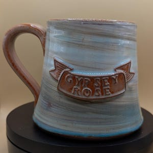 May include: Handmade ceramic mug with a brown handle and a light blue and brown exterior. The mug features a brown banner with the words "GYPSEY ROSE". The mug is a unique piece of drinkware.