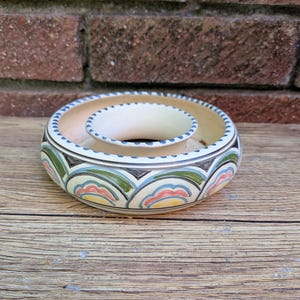May include: A round ceramic bowl with a central raised circular opening. The exterior features a repeating pattern of scalloped arches in shades of green, pink, yellow, and blue, outlined in black, with a matching dotted border along the top rim.