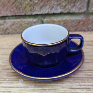 Op de afbeelding: Een kobaltblauwe kop en schotel set met gouden rand. De kop heeft een oor en een witte binnenkant. De schotel heeft dezelfde blauwe kleur met een gouden rand. De set staat op een houten oppervlak.