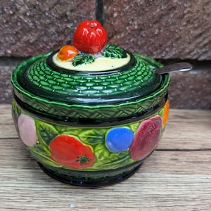 Könnte beinhalten: Eine Zuckerdose aus Keramik mit Deckel und kleinem Löffel. Die Schüssel ist grün mit einer gewebten Textur und mit bunten Gemüsesorten verziert. Der Deckel ist mit einer roten Tomate gekrönt. Die Schüssel steht auf einer Holzoberfläche.