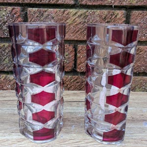 May include: Two clear drinking glasses with a geometric design. The glasses feature a pattern of red diamond shapes and clear glass. The glasses are cylindrical and sit on a wooden surface.