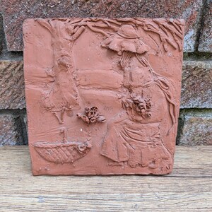 May include: A rectangular terracotta clay wall plaque featuring a relief sculpture of a figure wearing a wide-brimmed hat, sitting under a tree. The scene includes a woven basket, a small bouquet of flowers, and the initials T.N. etched in the corner.