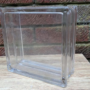 May include: A clear glass block with a rectangular shape. The block is transparent, allowing light to pass through. It is sitting on a wooden surface in front of a brick wall.