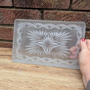 May include: A rectangular, frosted glass tray with a detailed starburst and diamond pattern. The tray has a scalloped edge and is held by a hand with a red painted fingernail. The tray is on a wooden surface.