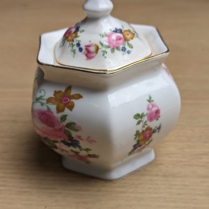 Puede incluir: Un azucarero de porcelana blanca con tapa, con un patrón floral en tonos de rosa, amarillo y azul. El tazón tiene una forma hexagonal y un borde dorado.