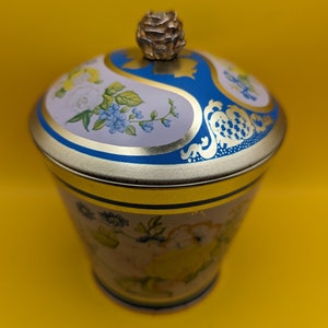 以下が含まれることがあります： 花柄の青い金色の蓋が付いたビンテージのブリキ容器。蓋には花の形をした装飾的なノブが付いています。容器は白い背景に花柄が描かれています。