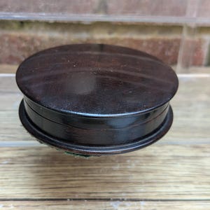 May include: A round, dark brown wooden box with a smooth, polished finish. The container features a flat, circular lid that sits flush with the base, creating a seamless appearance. The wood grain is subtle, and the edges are slightly rounded.