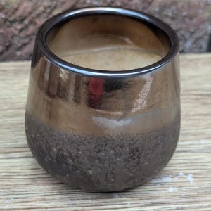 Può includere: Un bicchiere in ceramica marrone fatto a mano con una base strutturata unica. La tazza ha una finitura lucida e metallica che passa da un bordo marrone scuro a una sezione centrale marrone più chiara. L'interno è di colore beige chiaro.