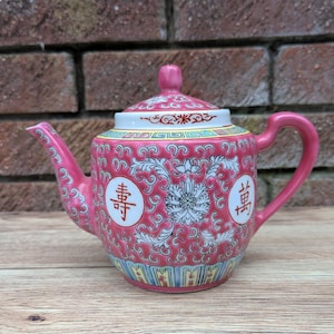 Peut inclure: Une théière en porcelaine rose avec un motif floral et des caractères chinois. La théière a un couvercle et une poignée.