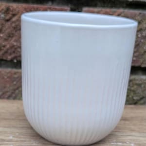 Może przedstawiać: Biały ceramiczny kubek z pionowymi żebrowanymi detalami. Kubek ma zaokrągloną podstawę i gładką, lekko rozszerzaną krawędź. Kubek stoi na drewnianej powierzchni.