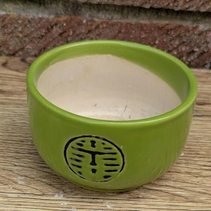 May include: A small, round, lime green ceramic bowl with a cream-colored interior. The bowl features a black, stylized symbol on the exterior. The bowl is sitting on a wooden surface.