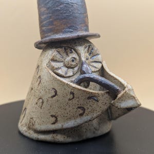 May include: A ceramic owl figurine wearing a brown top hat. The owl has a white and brown speckled body with a brown beak and eyes. The owl is holding a brown pipe in its beak.