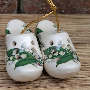 May include: A pair of miniature ceramic clogs, painted white with a floral design of green leaves and white flowers. The clogs have a gold cord for hanging. Decorative item.