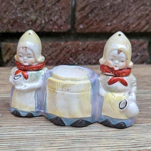 May include: A vintage ceramic planter featuring two Dutch girl figurines flanking a central pot. The figurines wear white bonnets, red scarves, and yellow aprons. The planter has a cream, blue, and yellow color scheme, with a distressed finish.