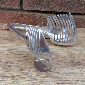 Könnte beinhalten: Ein silberfarbener Vintage-Tomatenschneider mit mehreren parallelen Klingen. Der Schneider ist so konzipiert, dass er Tomaten in gleichmäßige Scheiben schneidet. Der Schneider liegt auf einer Holzoberfläche.