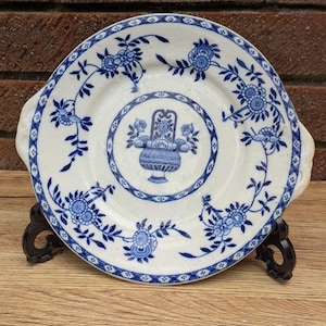 Puede incluir: Un plato de cerámica blanca con diseños florales azules. El plato tiene dos asas y un diseño central de una cesta de flores.