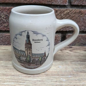 Peut inclure: Un chope à bière en céramique beige avec une anse, ornée d'une illustration détaillée du Rathaus de Hambourg. La chope a un marquage de capacité de 0,5L. Le fond est un mur de briques et une surface en bois.