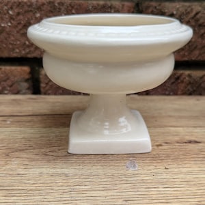 Może przedstawiać: Mała, kremowa ceramiczna miska na cokole z kwadratową podstawą, zaokrągloną środkową częścią i płytkim, okrągłym wierzchem. Obrzeże miski ma dekoracyjny, żebrowany wzór. Misa stoi na drewnianej powierzchni.