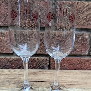 May include: Two clear glass champagne flutes featuring a gold-rimmed edge and a decorative design of red roses with the number 40 printed in red. Each flute has a tall, slender bowl and a vertical ribbed stem leading to a circular base.
