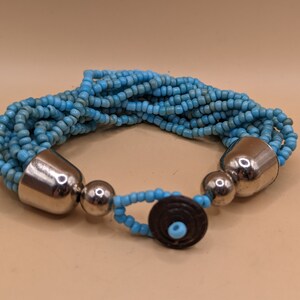 Op de afbeelding: Een meerstrengige armband gemaakt van lichtblauwe kralen met zilveren metalen accenten. De armband heeft een sluiting met een bruin houten midden.