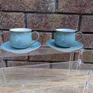 May include: Two matching ceramic tea cups and saucers in a soft, muted blue-green glaze. Each cup features a rounded, bulbous body with a curved handle and a smooth rim. The saucers are circular with a slightly raised center to hold the cup securely.