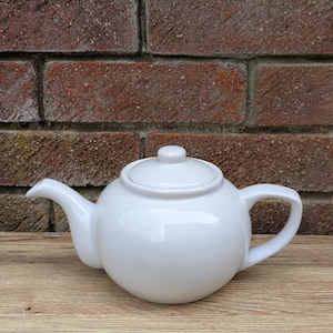 Peut inclure: Une théière en céramique blanche avec un corps rond et un couvercle. La théière a un bec verseur et une poignée.