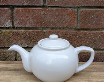 May include: A white ceramic teapot with a round body and a lid. The teapot has a spout and a handle.