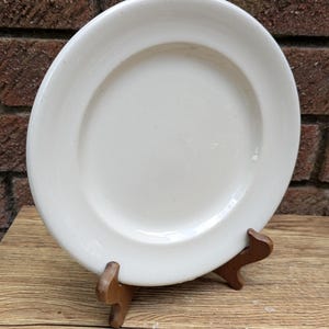 Può includere: Un piatto in ceramica bianca con un bordo leggermente rialzato, esposto su un supporto di legno. Il piatto ha un design semplice e classico ed è posto su una superficie di legno rustica e uno sfondo di mattoni.